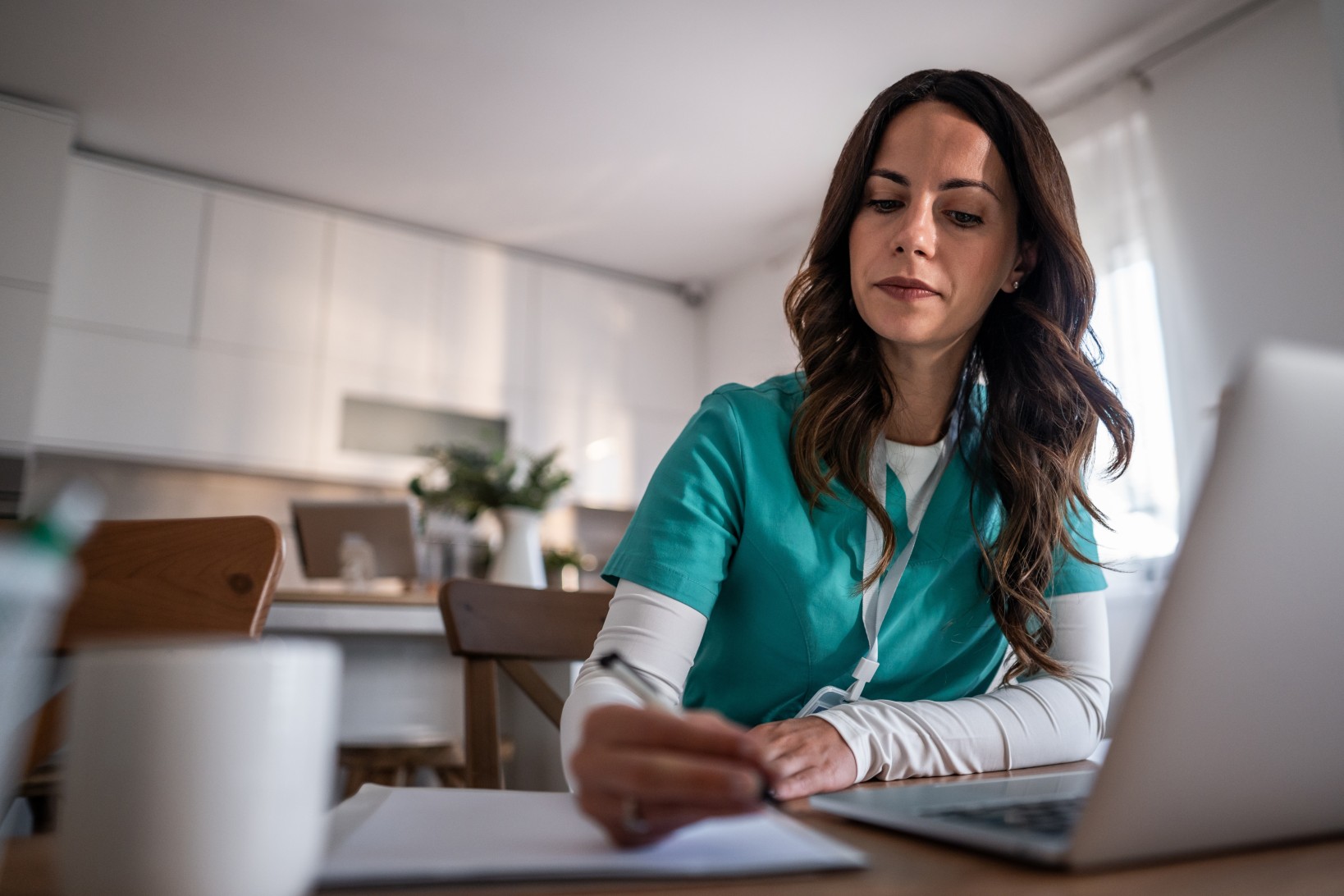 A woman in a teal medical scrubs is sitting at a table, writing on a notepad, with a laptop in front of her.